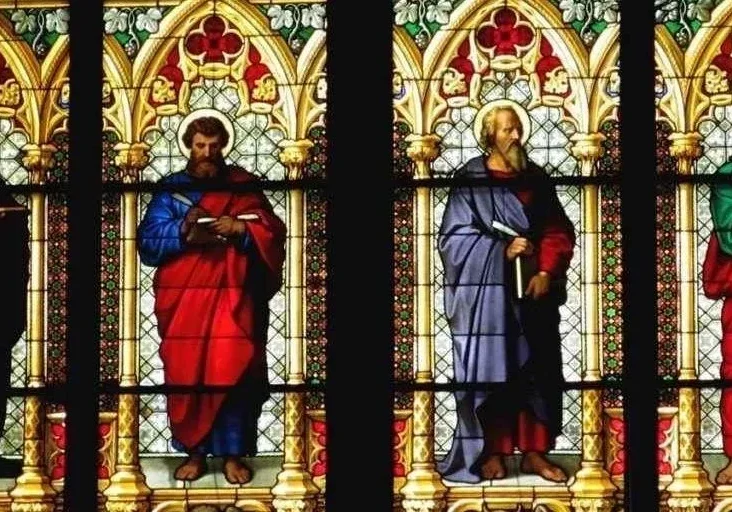 Stained glass window depicting four figures holding books and scrolls, dressed in red, blue, green, and purple robes, framed by ornate gothic arches.